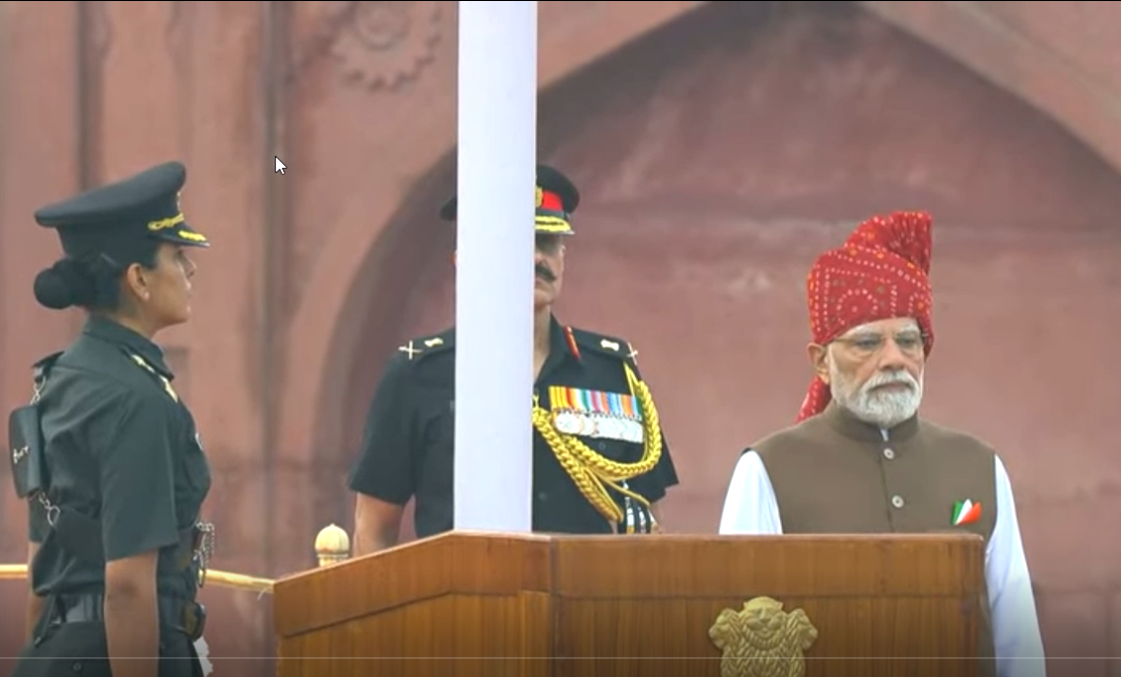 This image shows Prime Minister Narendra Modi standing at a wooden podium during the Independence Day ceremony at the Red Fort in New Delhi. He is wearing a brown sleeveless jacket over a white shirt and a distinctive red patterned turban. A small Indian flag pin is visible on his jacket. To his left stands a woman officer in a dark uniform with a peaked cap. Behind them is a senior military officer in ceremonial uniform with medals. A white flagpole is positioned between them. The background features the red sandstone walls and arched architecture of the Red Fort. The scene is from the 15 August 2026 Independence Day event.
